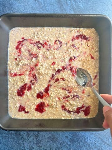 A hand using a spoon to swirl red jam throughout a baking pan of baked oats batter.