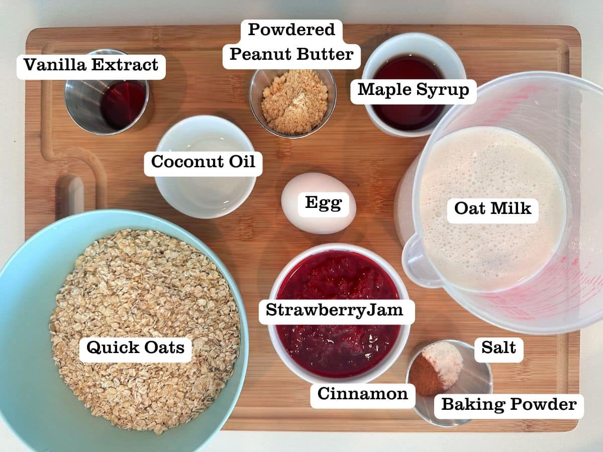 A wooden board containing ingredients for PB and J baked oats, including powdered peanut butter, strawberry jam, oat milk, and maple syrup.