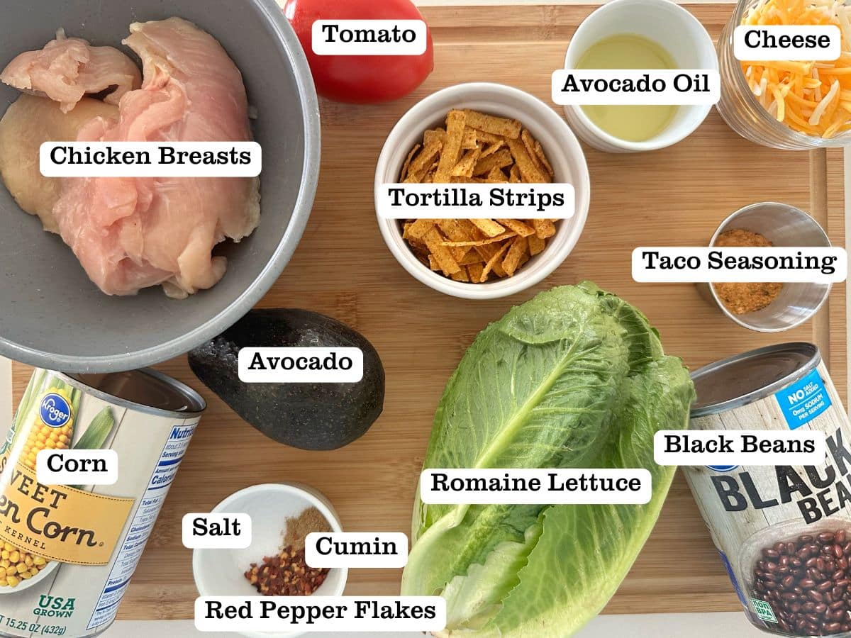A wooden board containing ingredients for southwest chicken salad, including black beans, tortilla strips, and avocado.