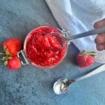 A jar containing homemade strawberry preserves with a spoon scooping up a portion.