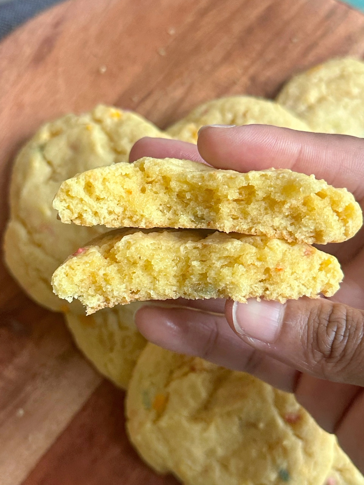 A hand holding a sprinkle cookie that has been cut in half to display the texture inside.