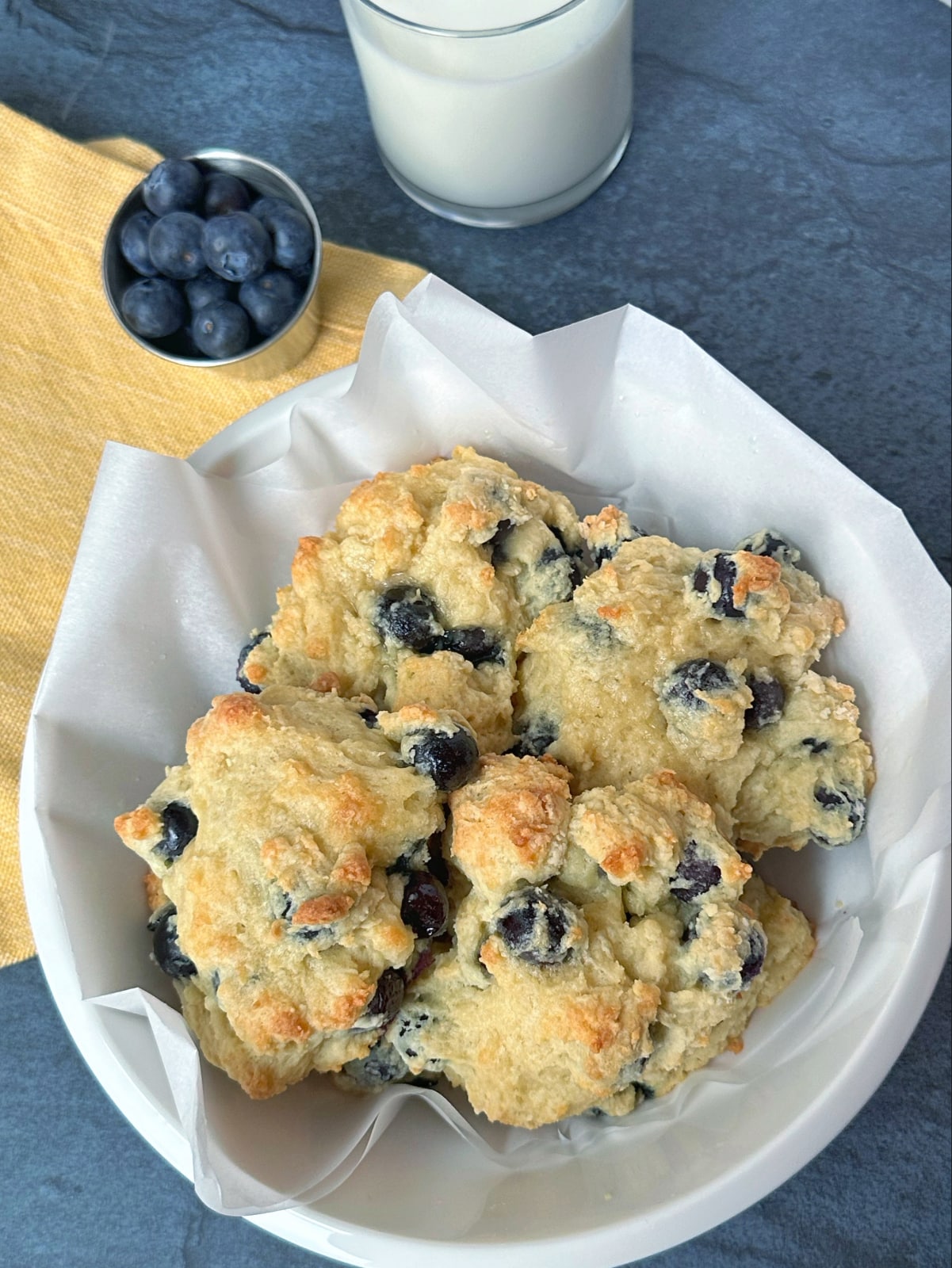 A white bowl filled with blueberry biscuits with a glass of milk and cup of blueberries on the side.