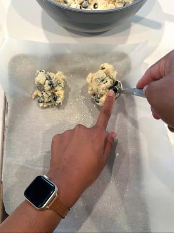 A pair of hands using a spoon to drop blueberry biscuit dough onto a parchment-lined sheet pan.
