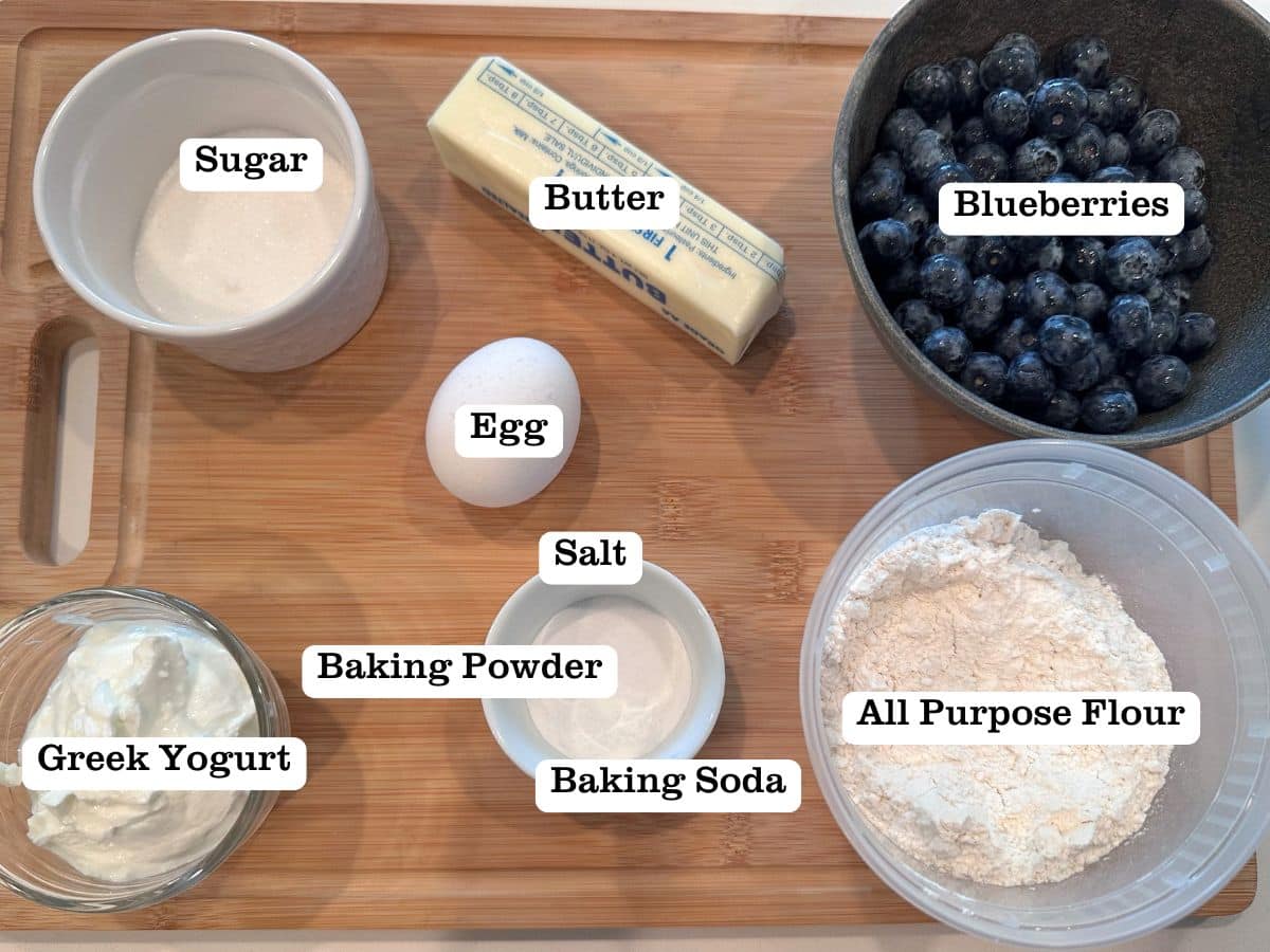 A wooden board containing ingredients for blueberry drop biscuits, including blueberries, butter, sugar, and Greek yogurt.