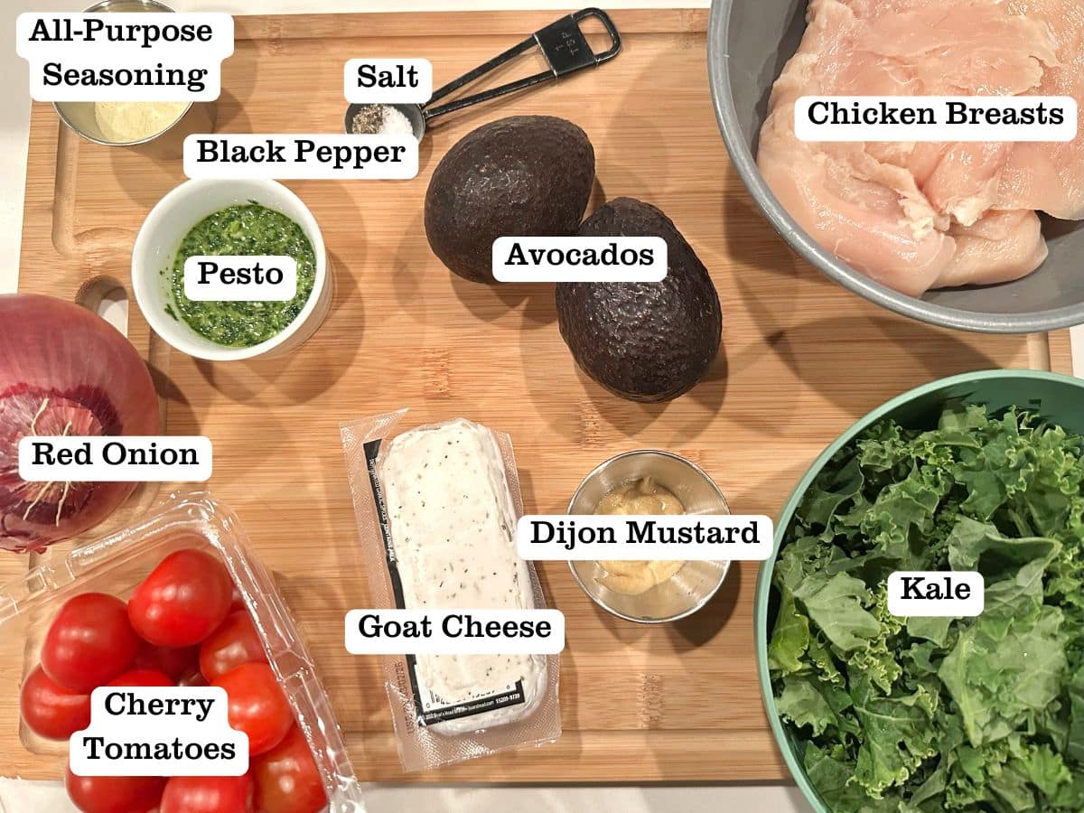 A wooden board containing ingredients for a salad, including pesto, chicken breasts, goat cheese, and avocados.