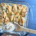 A spoon scooping out a portion of seafood stuffed shells in a homemade cream sauce in a glass baking dish.