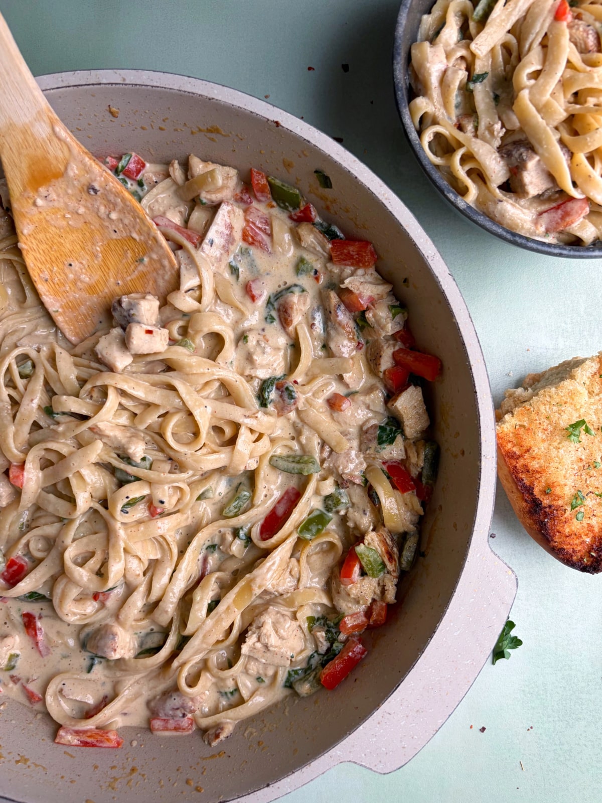 A skillet of Jerk Chicken Alfredo with a wooden serving spoon sticking out of it, next to a piece of garlic bread.