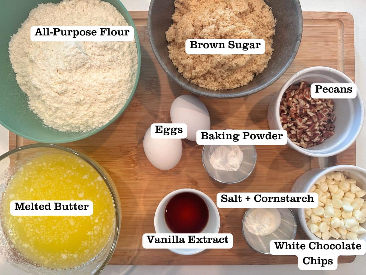 A wooden board containing the ingredients for brown sugar blondies, including butter, flour, chopped pecans, and white chocolate chips.
