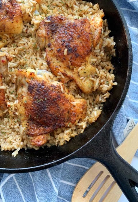 A cast iron skillet filled with One Skillet Chicken Thighs & Rice, with a wooden spoon on the side.