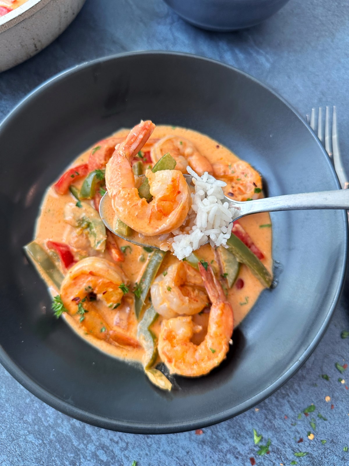 A spoon holding up a bite of rice and shrimp curry, over a black bowl filled with more soup.