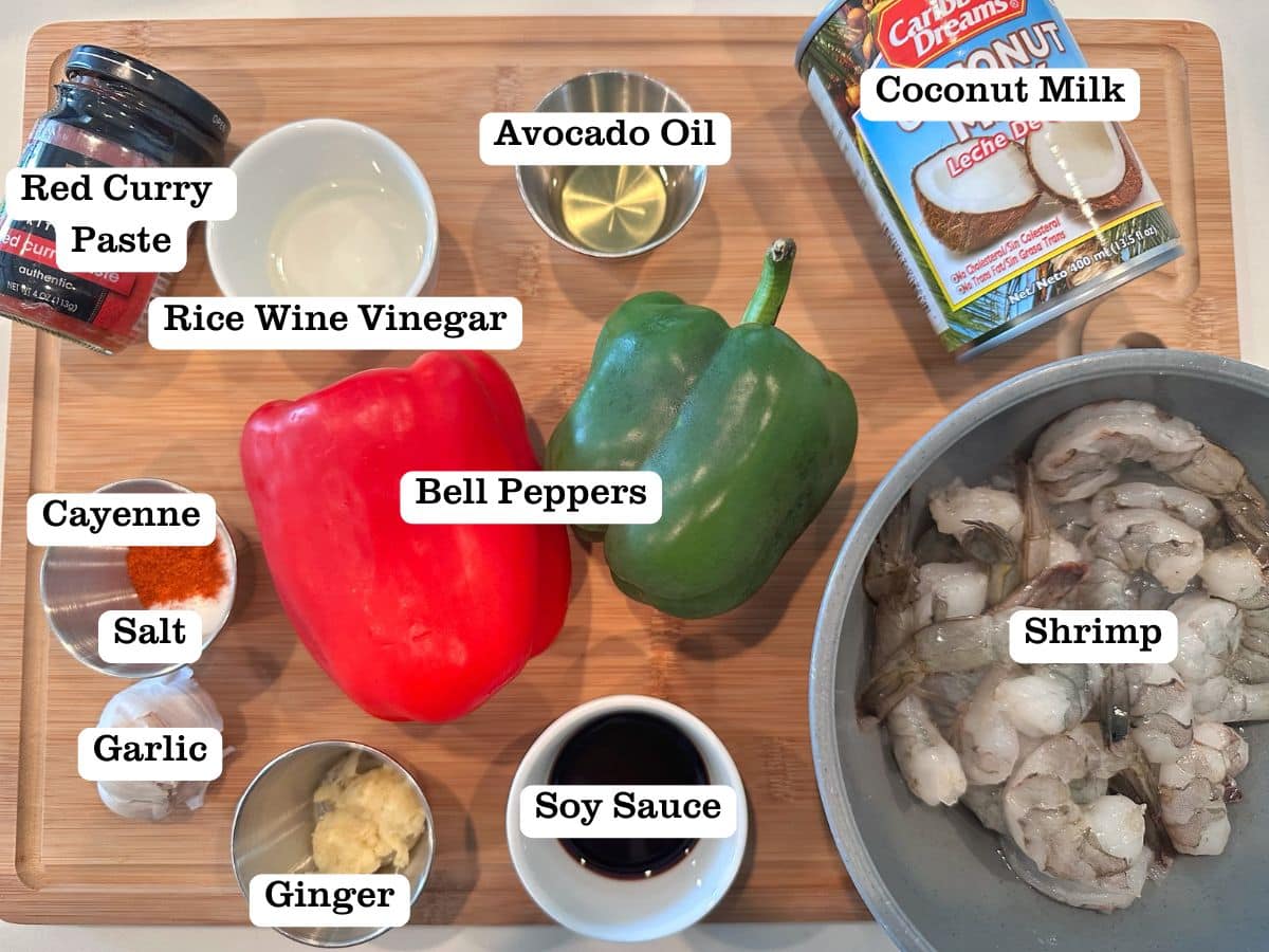A wooden board containing ingredients for red curry including curry paste, bell peppers, and a can of coconut milk.