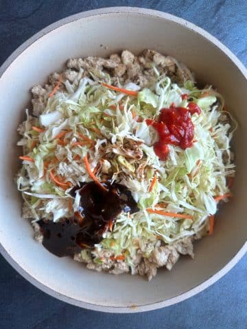 A pan containing cooked ground chicken, shredded cabbage and carrots, and brown and red sauces.