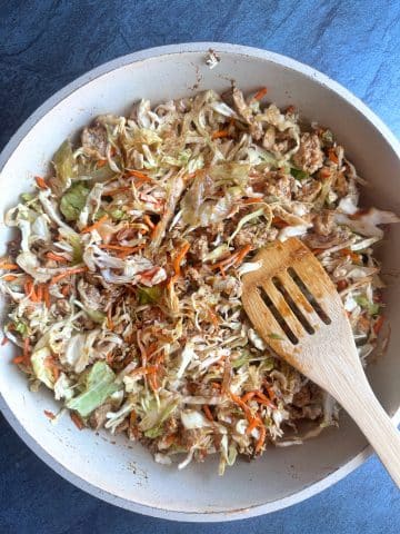 A cream colored pan containing mixed ground chicken and shredded vegetables with a wooden spoon sticking in it.