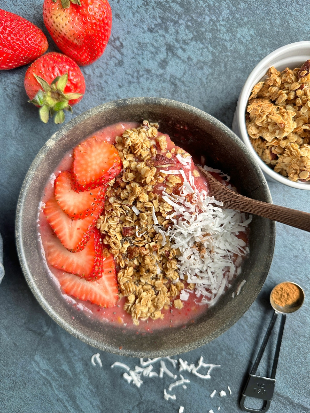 A spoon sticking out of the side of a pink smoothie bowl topped with berries, granola, and coconut.