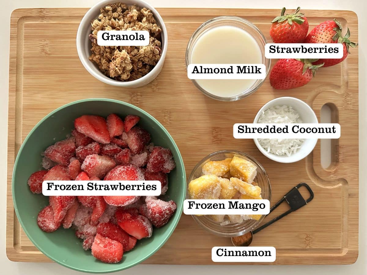 A wooden board containing bowls of ingredients for a strawberry-mango smoothie bowl.