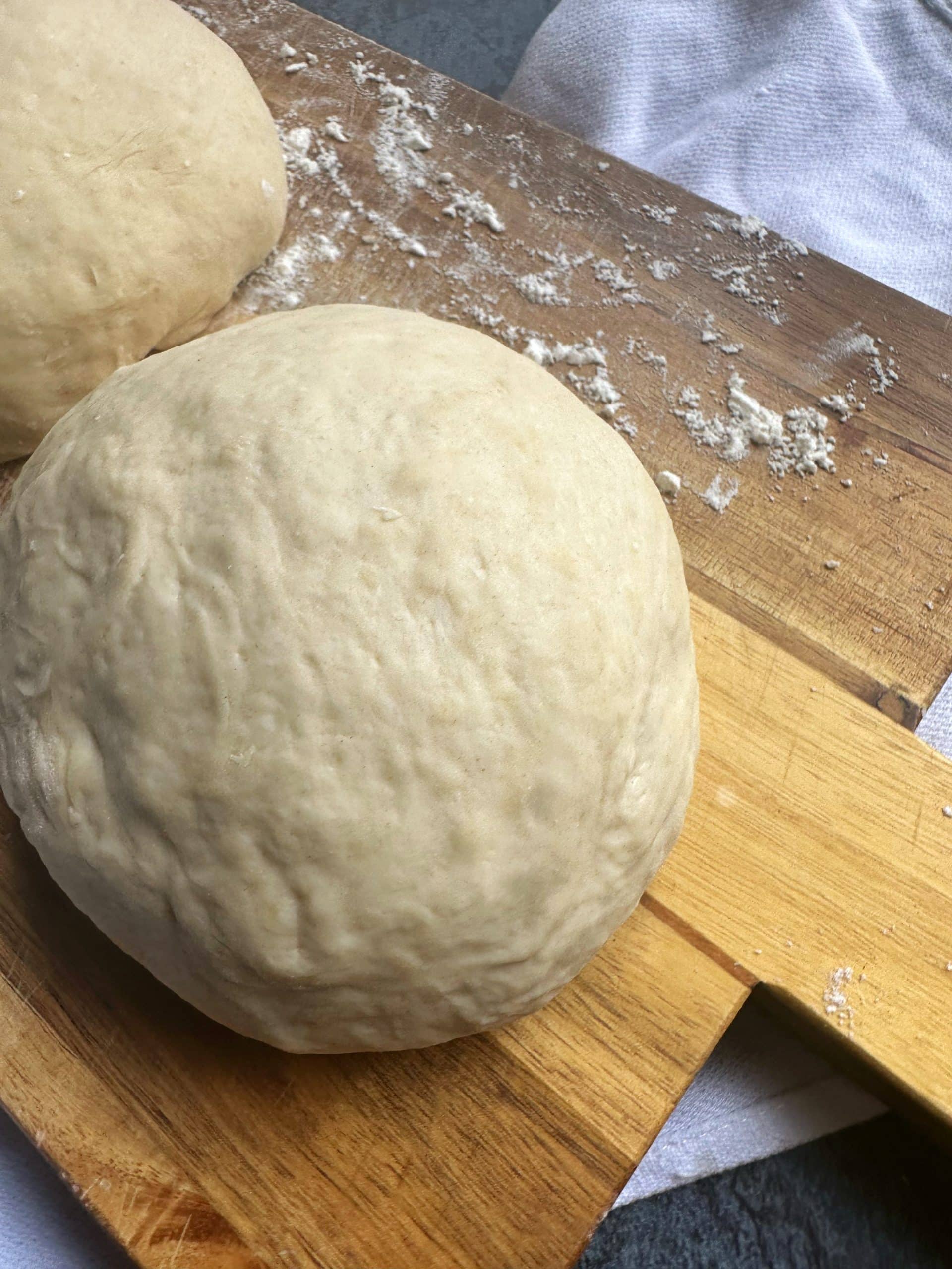 A flour-dusted wooden board containing two balls of no-rise homemade pizza dough with yeast.