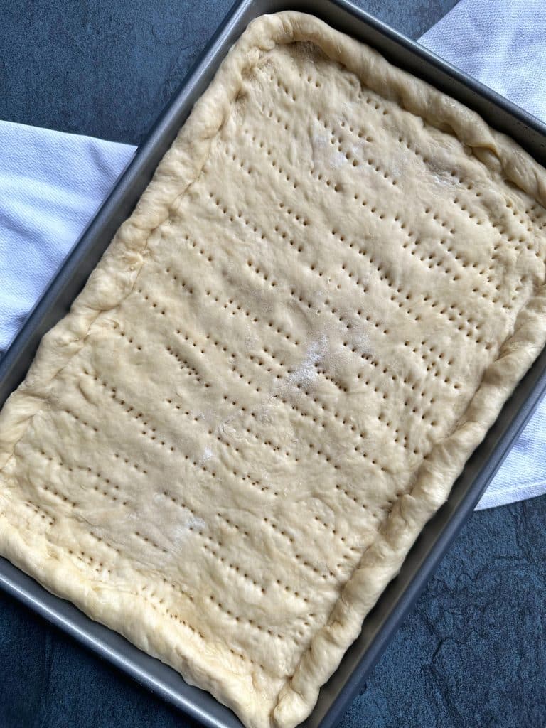 A tray of no-rise homemade pizza dough, shaped into a rectangle, with fork holes pierced throughout.