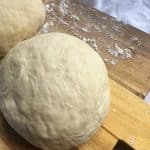 A flour-dusted wooden board containing two balls of no-rise homemade pizza dough with yeast.