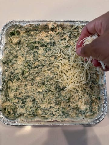 A hand sprinkling white shredded cheese onto an aluminum pan filled with unbaked spinach dip.