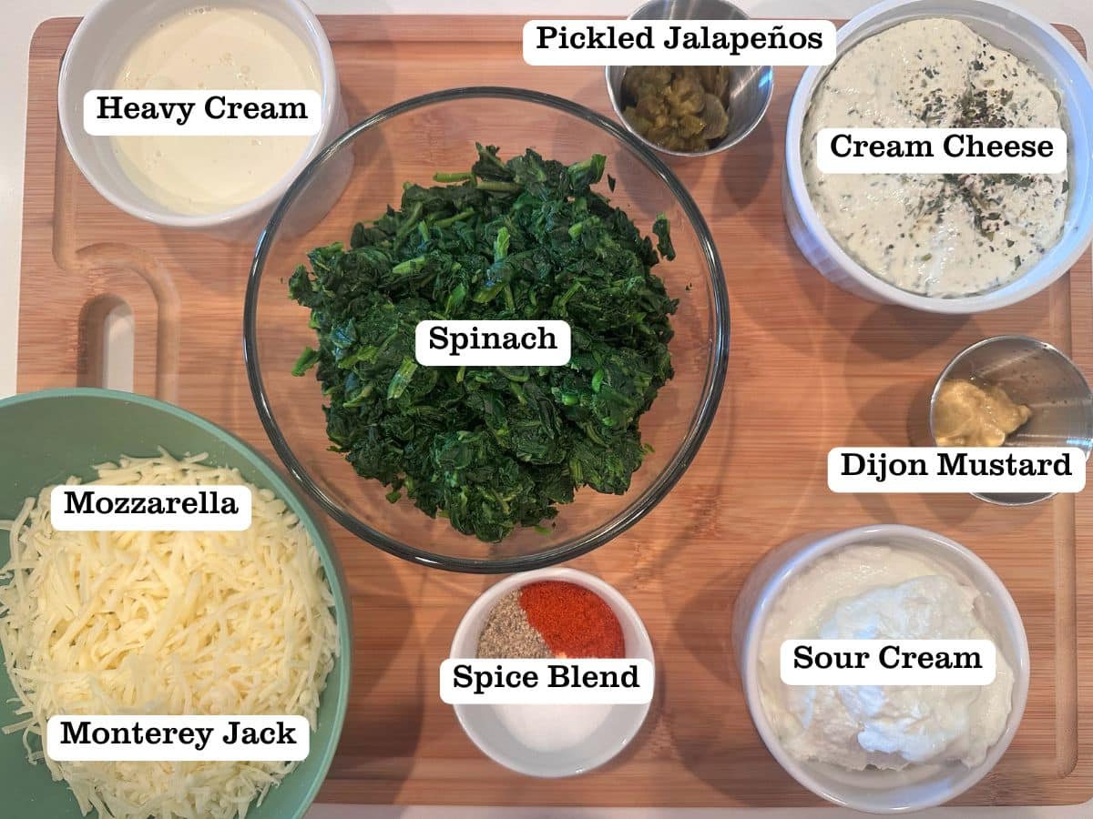 A wooden board containing ingredients for spinach dip, including Monterey Jack cheese, cream cheese, and pickled jalapeños. 