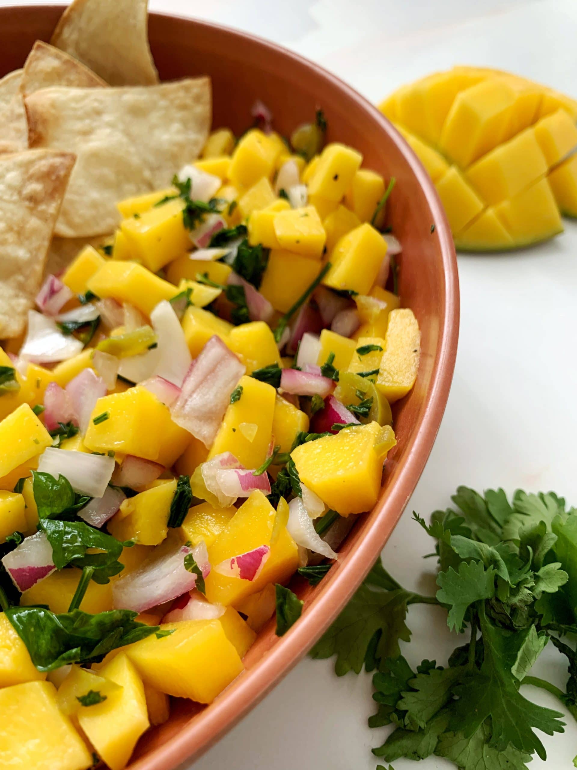 bowl of homemade mango salsa with tortilla chips, cilantro, and a scored mango on the side