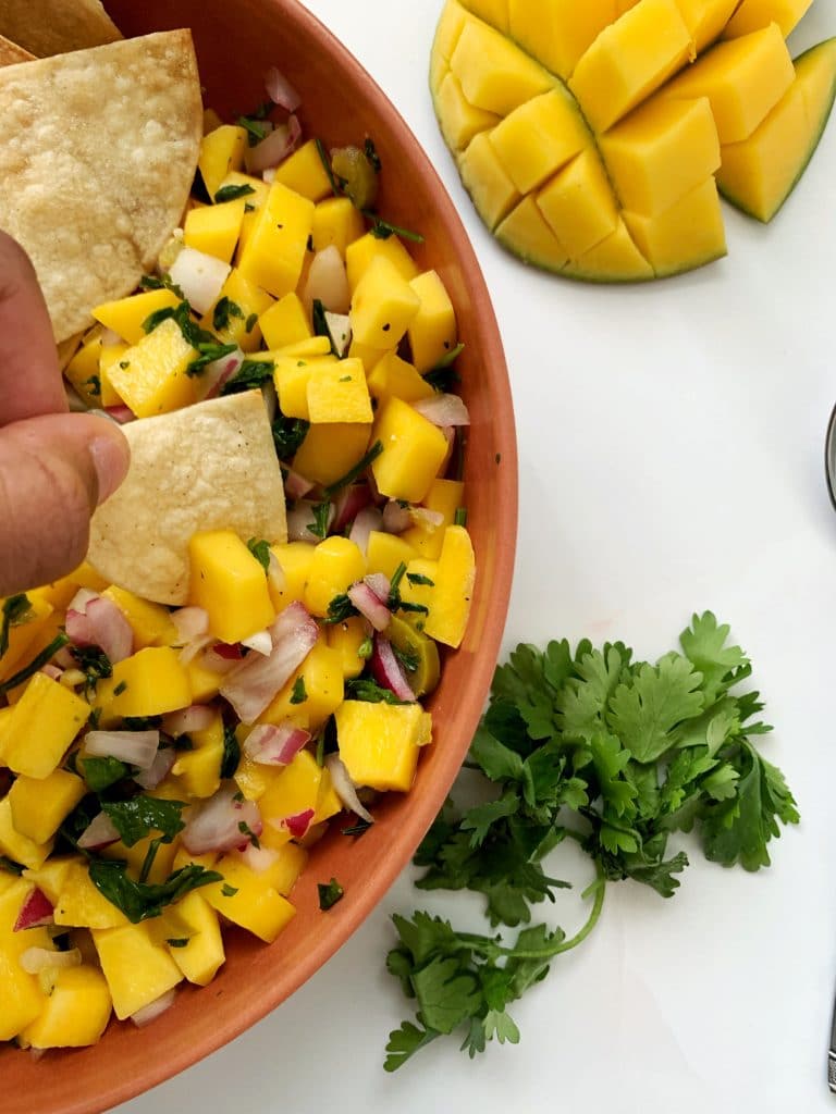 bowl of homemade mango salsa with tortilla chips, cilantro, and a scored mango on the side