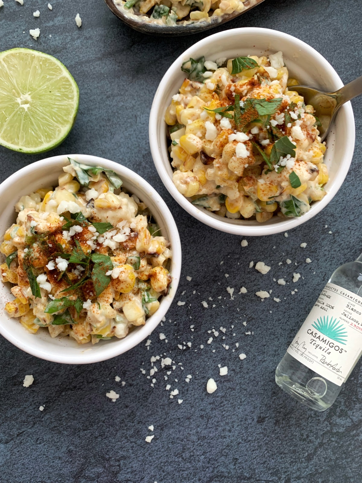 Two bowls of esquites (elote in a cup) with lime slices and a mini tequila bottle in the background.