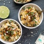 Two bowls of mexican corn salad (esquites) with tequila and a lime on the side.
