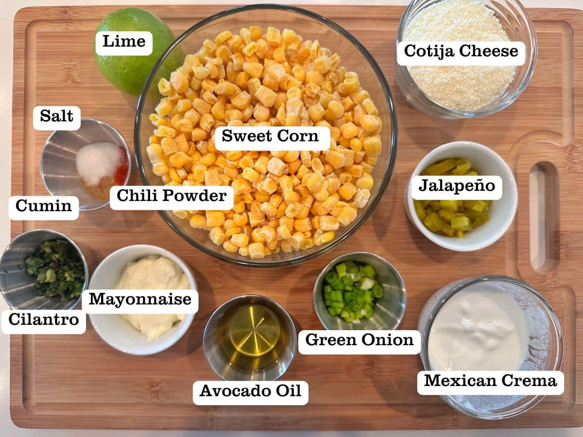 A wooden board containing ingredients for esquites, including corn, mayo, crema, and cotija cheese.