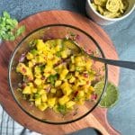 A glass bowl filled with homemade mango salsa on a wooden serving board.