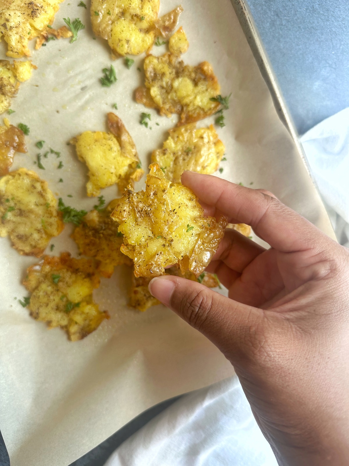 A hand holding a singular smashed potato, with the remaining ones on a sheet pan in the background.