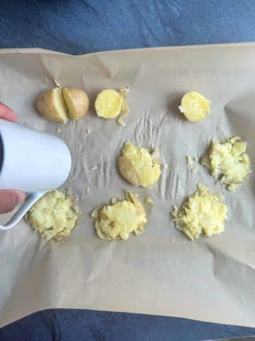 A hand using the bottom of a mug to smash boiled potatoes on a sheet pan before roasting.