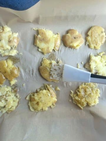 A hand brushing a seasoning and butter blend onto boiled, smashed potatoes.