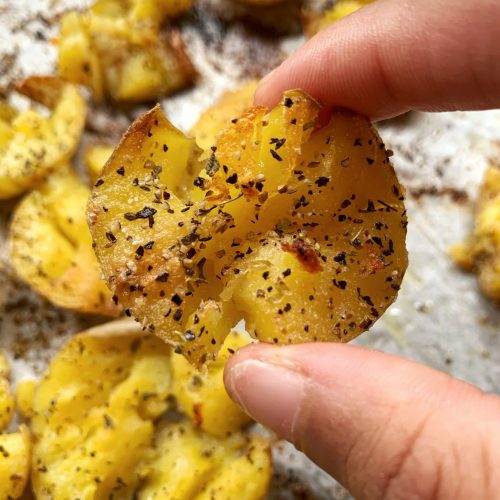 a finger holding a crispy smashed potato with more potatoes in the background on a sheet of crumbled parchment paper