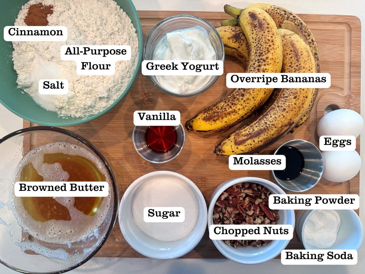 A wooden board containing ingredients for banana bread, including bananas, browned butter, sugar, nuts, and flour.