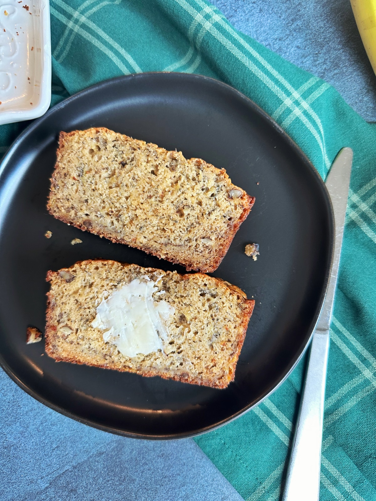 Two slices of brown butter banana bread on a black plate with a pat of butter in the middle of one slice.