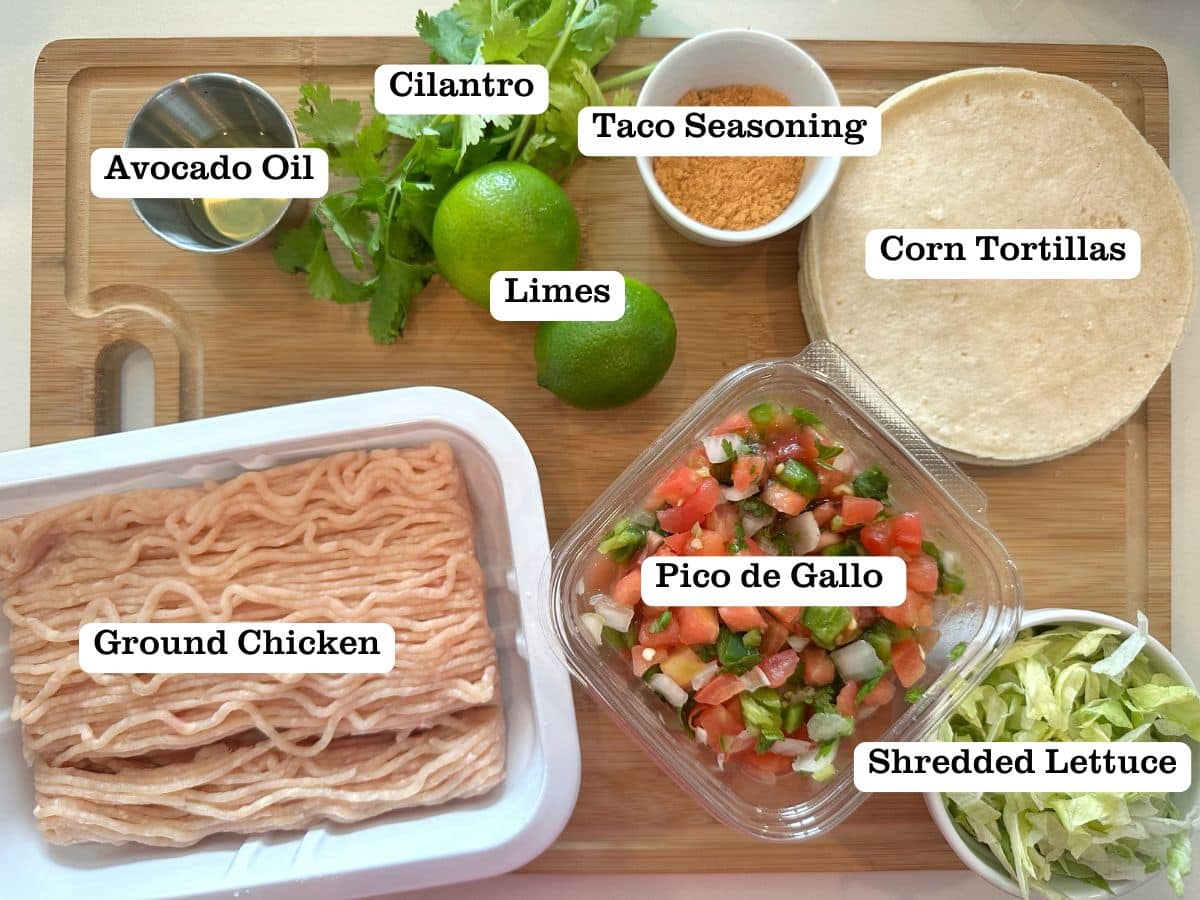 A wooden board containing ingredients for chicken tacos; including ground chicken, pico, limes, and corn tortillas.