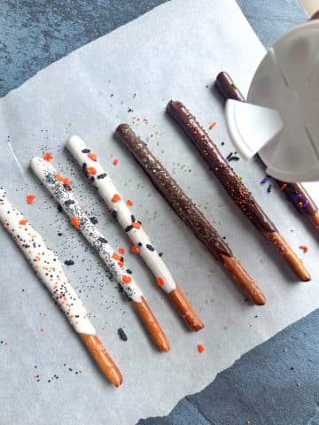 A hand sprinkling Halloween-themed sprinkles onto chocolate covered pretzel rods.