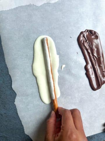 A hand rolling jumbo pretzel rods into white chocolate spread on a piece of parchment paper.
