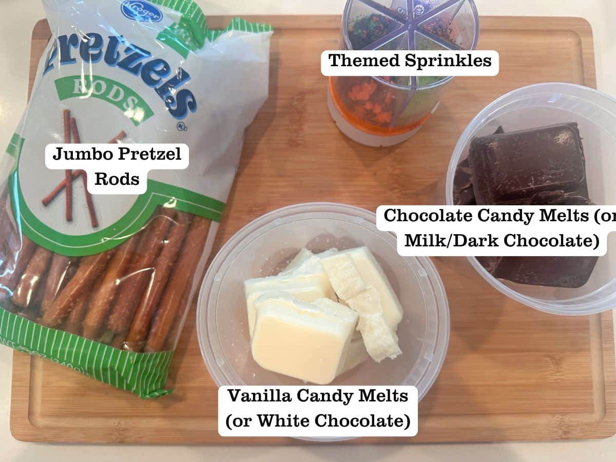A wooden board containing deli containers of white and milk chocolate candy melts, Halloween sprinkles, and pretzel rods.