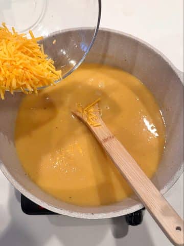 A bowl of sharp cheddar being poured into a dutch oven filled with soup.