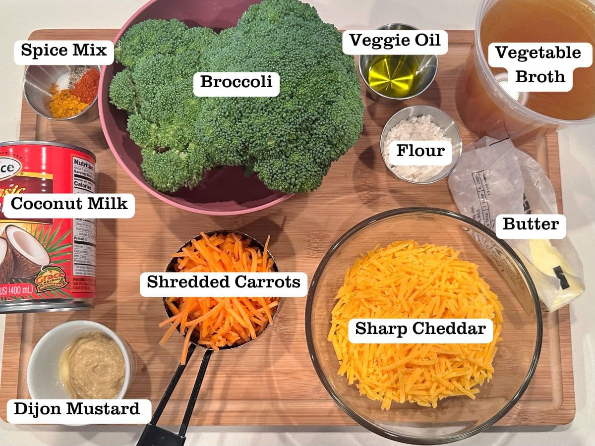 A wooden board with soup ingredients like sharp cheddar, broccoli, carrots, and coconut milk.