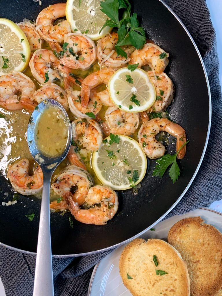 Skillet with garlic butter shrimp scampi, lemon slices, and parsley with bread on the side.