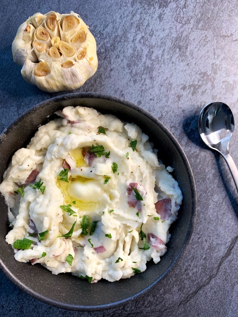 Roasted garlic red skin mashed potatoes in a black bowl with a head of roasted garlic on the side.