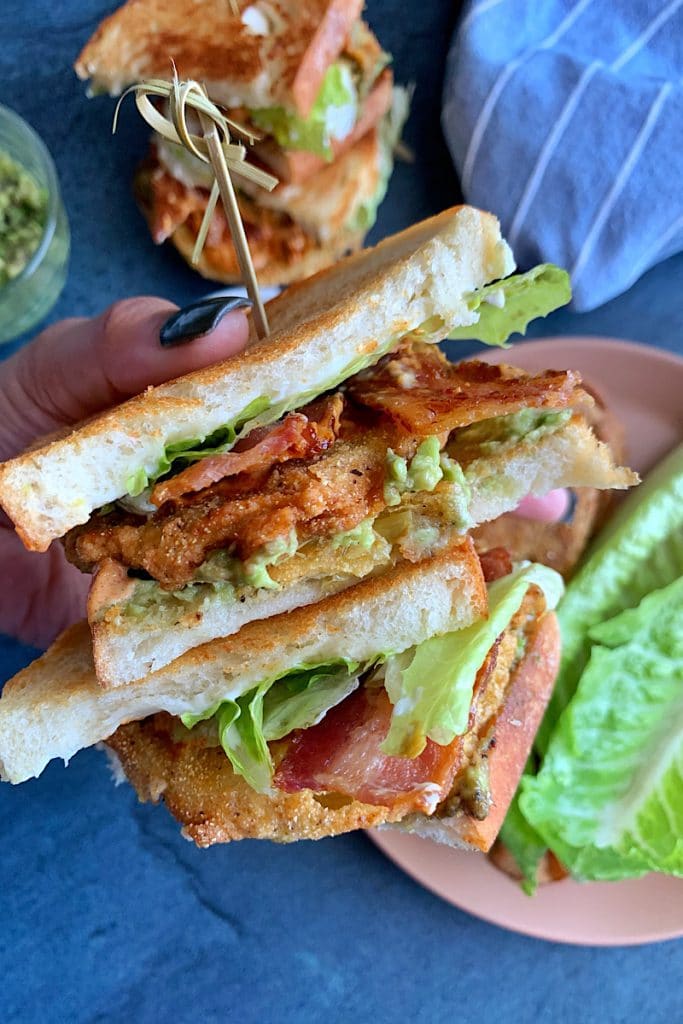 A hand holding two stacks of Fried Green Tomato sandwiches with more sandwich ingredients in the background.