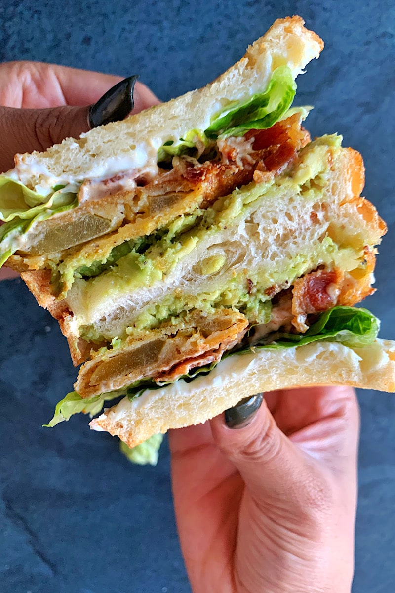Two hands holding a Fried Green Tomato BLT sandwich cut in half.