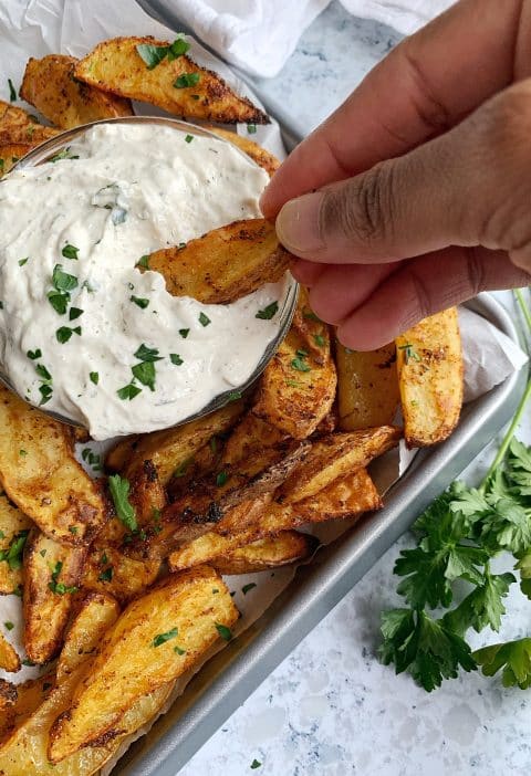 An Air Fried Potato Wedge Dipped into Homemade High-Protein Ranch with more ranch and potatoes on a tray in the background.