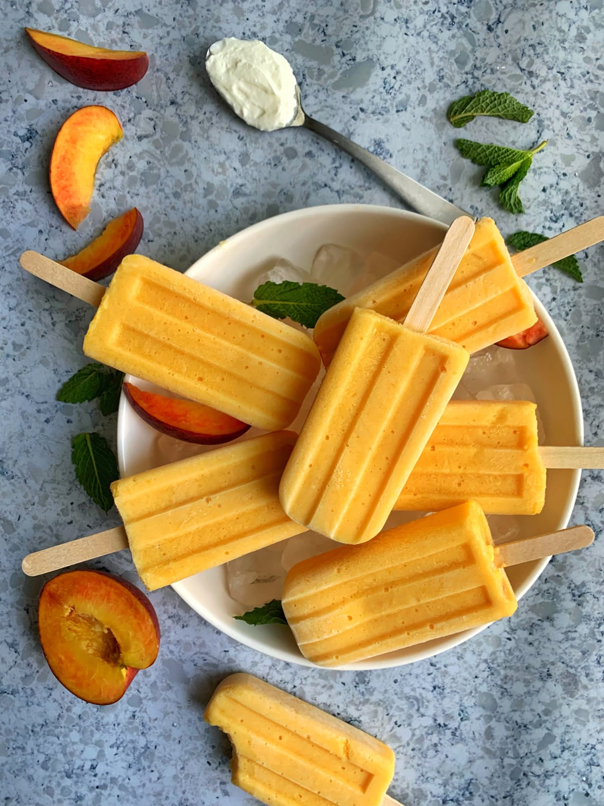 A bowl of ice covered with homemade peaches and cream popsicles, with extra ingredients placed in the background.