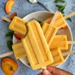 Homemade creamy peach popsicles on a bowl of ice with mint leaves, sliced peaches, and a spoon of greek yogurt in the background.