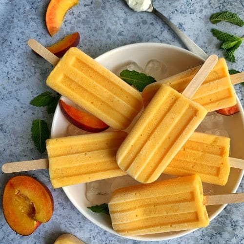 Homemade creamy peach popsicles on a bowl of ice with mint leaves, sliced peaches, and a spoon of greek yogurt in the background.